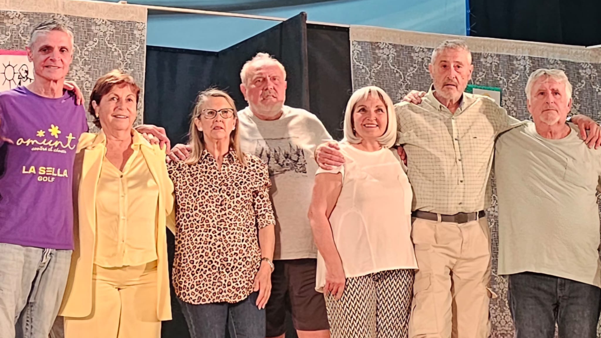 Un plano medio de seis personas del elenco y equipo técnico del teatro solidario "És molt important…" de Desficaci Teatre, posando abrazados y sonrientes frente al público. De izquierda a derecha: un hombre con camiseta morada de "Amunt", una mujer con traje amarillo, una mujer con blusa de leopardo y gafas, un hombre con camiseta gris con un gráfico de árboles, una mujer con blusa blanca y pantalones estampados, y un hombre con camisa a cuadros y pantalones cargo.
