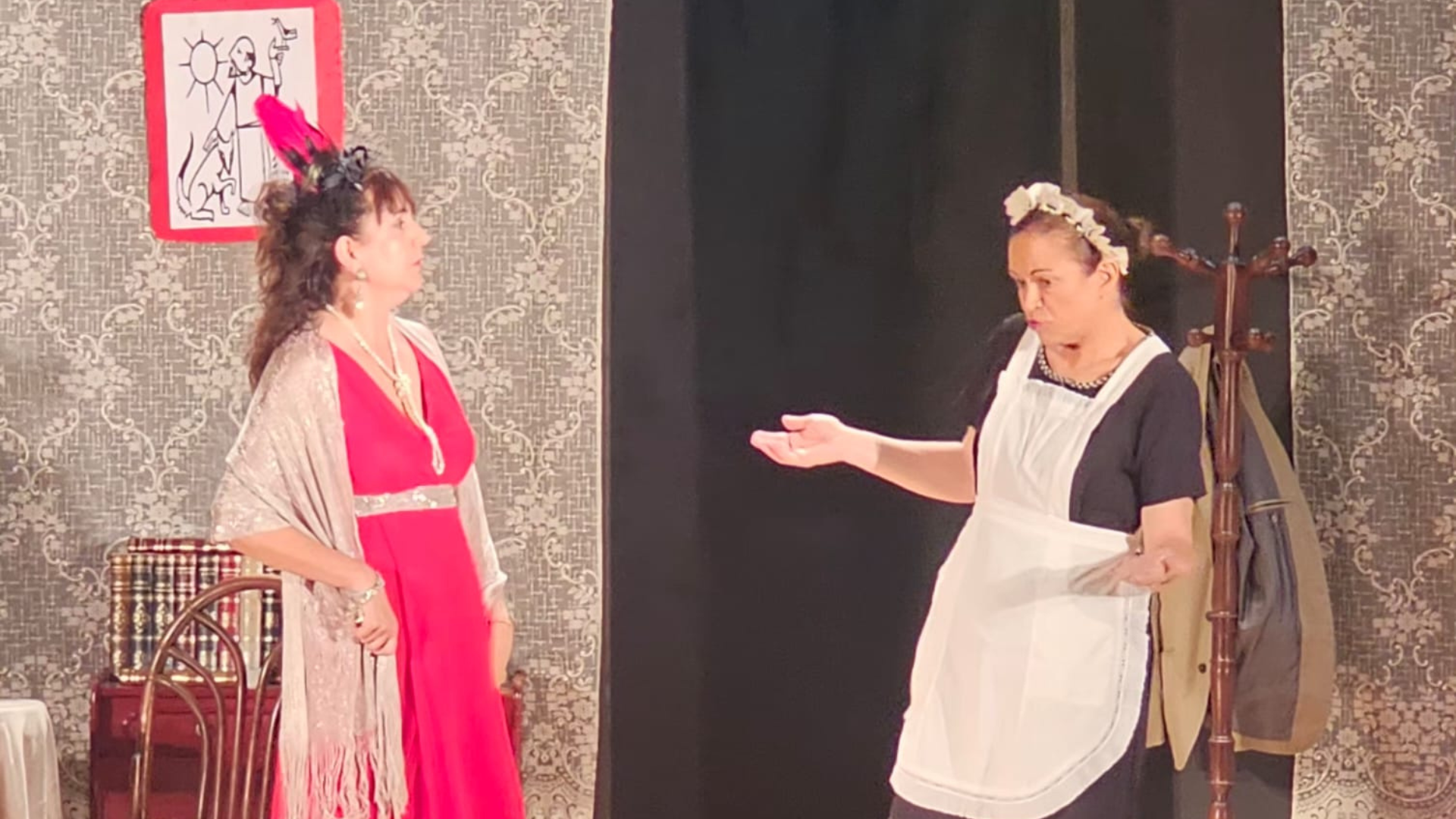 Dos actrices de Desficaci Teatre en una escena dramática de "És molt important… història d’un misteriós assassinat". Una mujer con un elegante vestido rosa fucsia y un tocado de plumas rojas mira hacia el público con expresión seria, mientras una sirvienta con uniforme negro y delantal blanco gesticula con una mano extendida. El escenario incluye un perchero y papel tapiz de fondo.