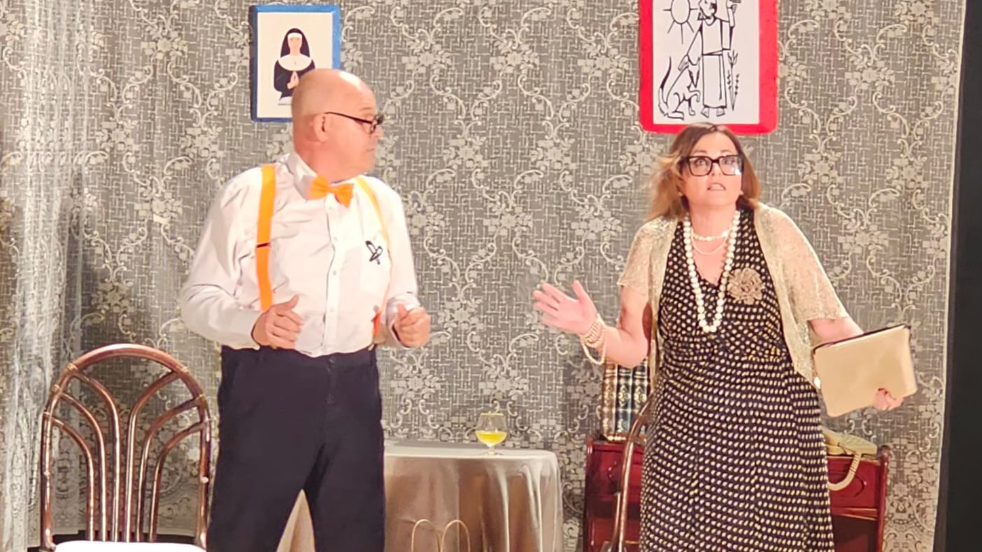 Dos actores de Desficaci Teatre en escena durante la obra solidaria "És molt important…". Un hombre con pajarita y tirantes naranjas gesticula frente a una mujer con un vestido de lunares y collar de perlas, quien parece sorprendida y sostiene un bolso de mano beige. El fondo muestra un decorado con papel tapiz y cuadros en el Auditori del CEIP Llebeig en Dénia.