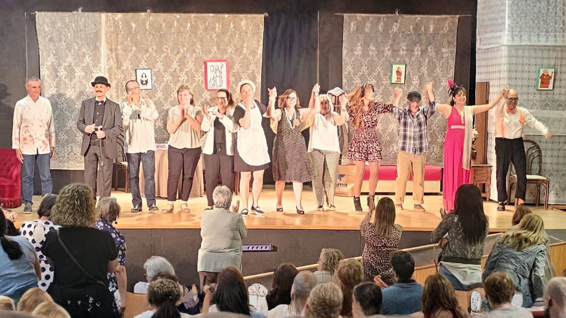 Vista general del elenco de Desficaci Teatre saludando de la mano al público al finalizar la obra benéfica "És molt important…" en el escenario del Auditori del CEIP Llebeig de Dénia. La sala está llena de espectadores aplaudiendo de pie y sentados. Varios actores visten disfraces, incluyendo una sirvienta, un hombre de negocios y personajes con vestidos de noche.