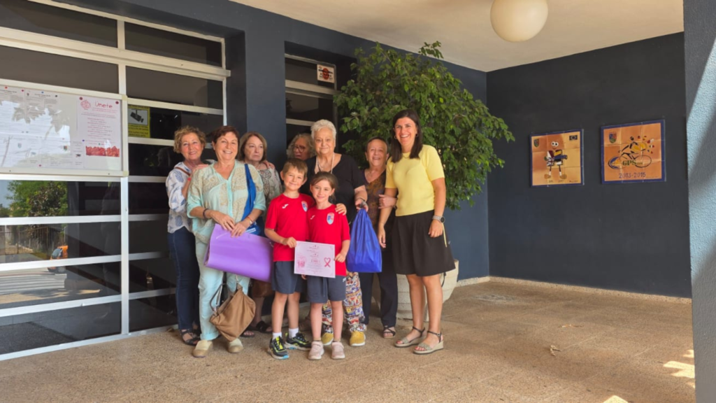 Un grupo de mujeres, una profesora y dos niños con el uniforme deportivo del Colegio Paidos posan sonrientes en la entrada del centro escolar, sosteniendo un cartel y material escolar.