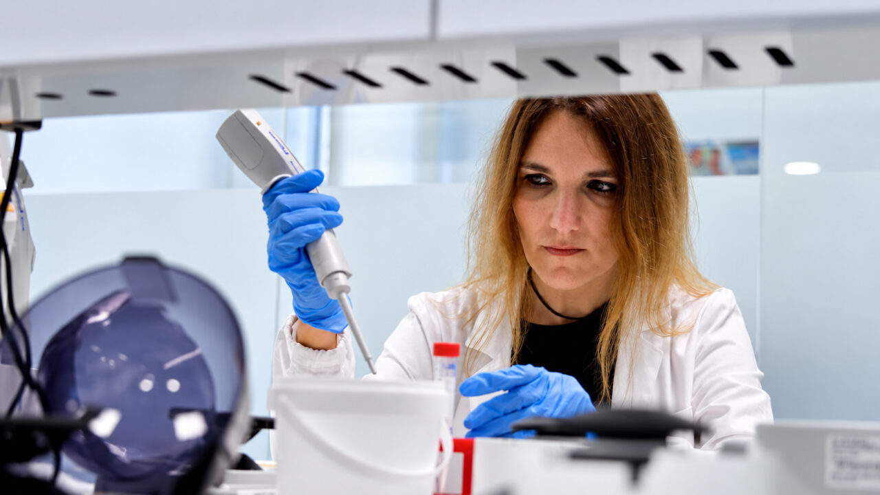 Noelia Tarazona, doctora del INCLIVA, trabajando en un laboratorio de investigación biomédica, concentrada mientras manipula muestras con una pipeta de precisión y lleva guantes azules. Ilustra la noticia del blog de Amunt sobre biopsia líquida.