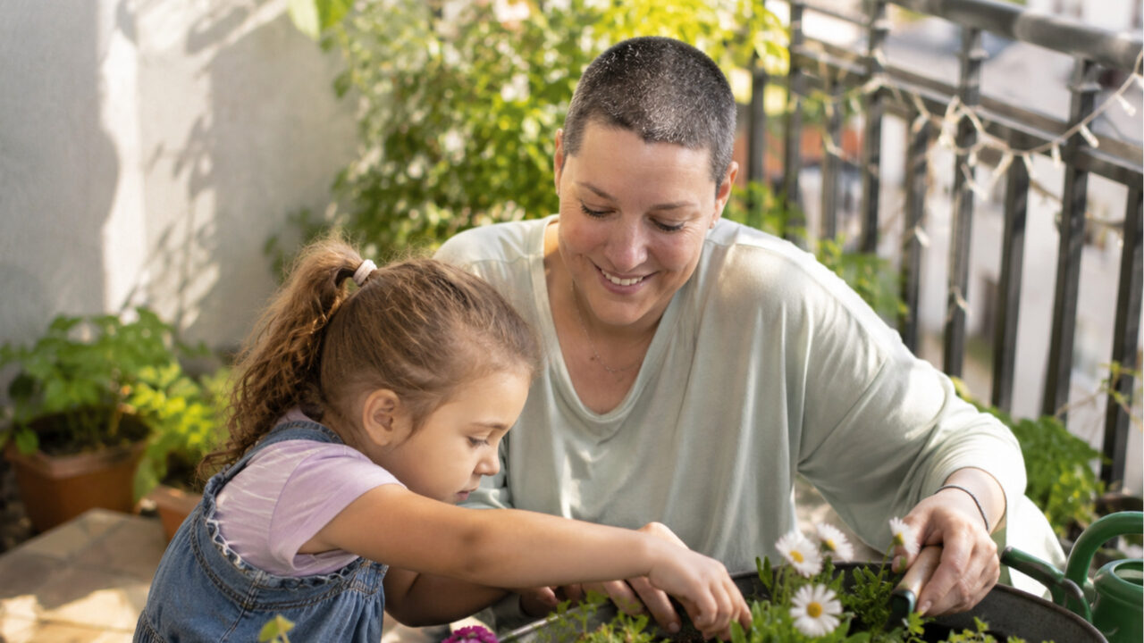 Una mujer con la cabeza rapada y una niña comparten un momento feliz de jardinería en un balcón soleado, un ejemplo de calidad de vida