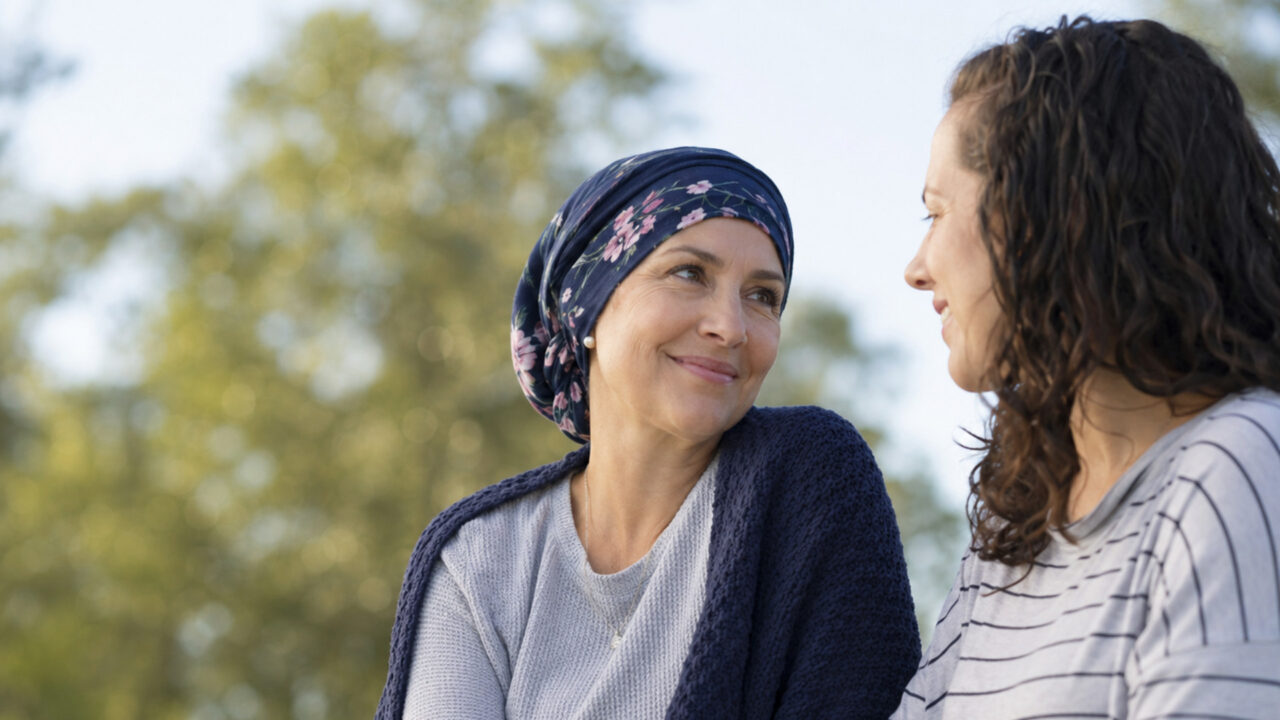 Dos mujeres adultas conversan en el exterior. Una lleva un pañuelo en la cabeza y mira con gratitud a su amiga. Esta conexión emocional es clave para su calidad de vida.