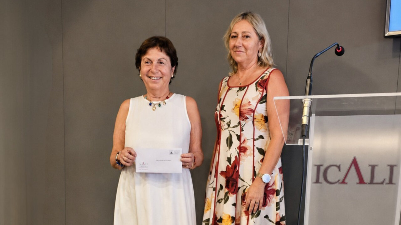 Dos mujeres posan sonrientes en un estrado durante la entrega de una ayuda institucional. La mujer de la izquierda, con un vestido blanco, sostiene un sobre con el logotipo de ICALI. A su lado, la segunda mujer luce un vestido con estampado floral. En primer plano, a la derecha, destaca un atril de metacrilato donde se lee claramente la inscripción ICALI.