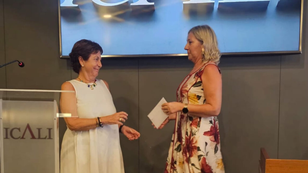 Momento de la entrega oficial de la subvención en la sede del Colegio de Abogados. Una mujer con un vestido de flores entrega un sobre blanco a otra mujer vestida de blanco, quien lo recibe con una sonrisa cerca del atril con el logotipo de ICALI. Al fondo se observa una gran pantalla de televisión instalada en una pared gris.