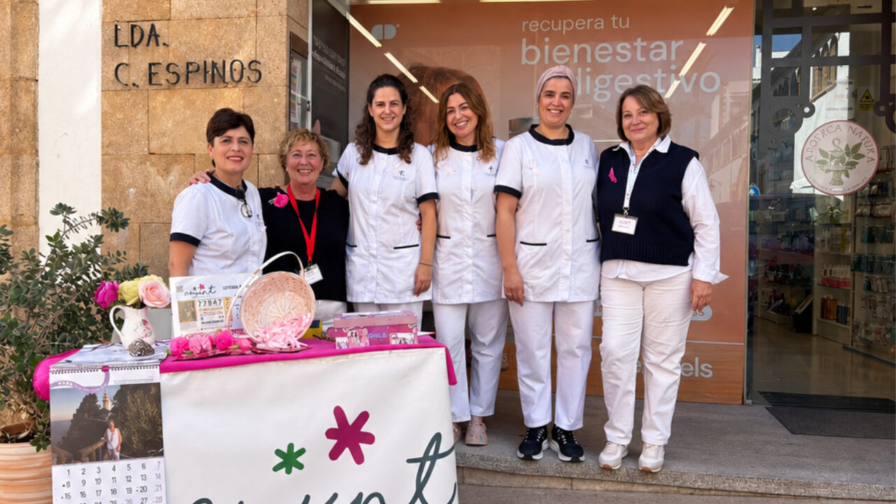 Texto Alt: Seis mujeres, entre personal sanitario con uniforme blanco y voluntarias de Amunt contra el Càncer que también participan en la Feria de Asociaciones de Dénia, posan en la entrada de la Farmacia Mar Espinós en Xàbia junto a una mesa con lazos rosas y folletos informativos sobre la prevención del cáncer de mama.