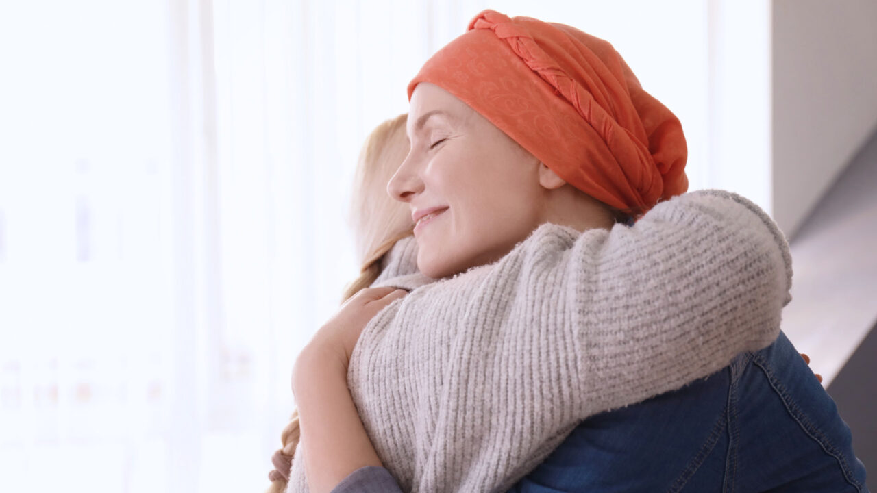Primer plano de una mujer sonriente con un pañuelo oncológico naranja abrazando a otra persona por la espalda, con los ojos cerrados y expresión de paz. La imagen transmite el apoyo y la esperanza que representa la campaña "Loteria Solidaria Amunt 2026", con luz natural entrando por una ventana lateral.